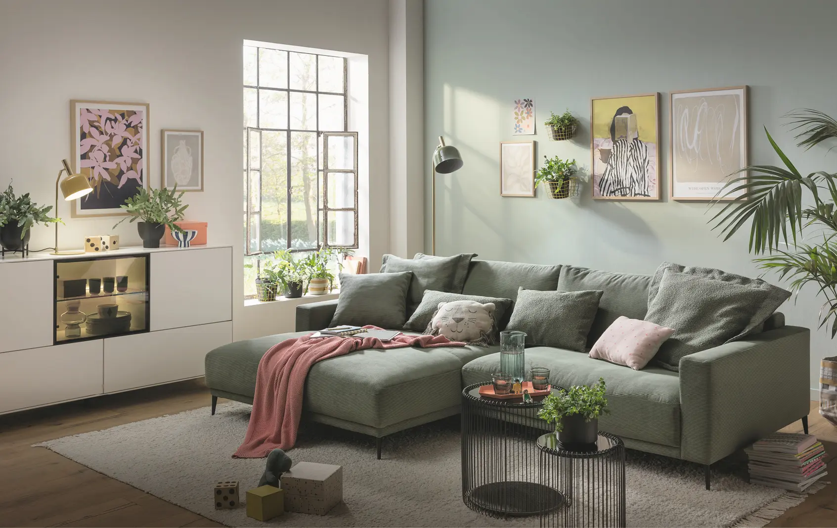 Wohnzimmer mit grünem Ecksofa, Kissen und Decke, davor runder Couchtisch und Teppich, links Sideboard mit Pflanzen